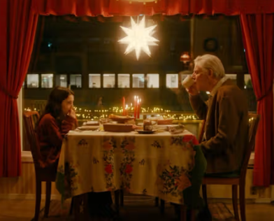 Bildet av ei jente og en mann som sitter rundt et bord fullt av mat. I bakgrunnen er det et vindu med en julestjerne og røde gardiner. - Klikk for stort bilde