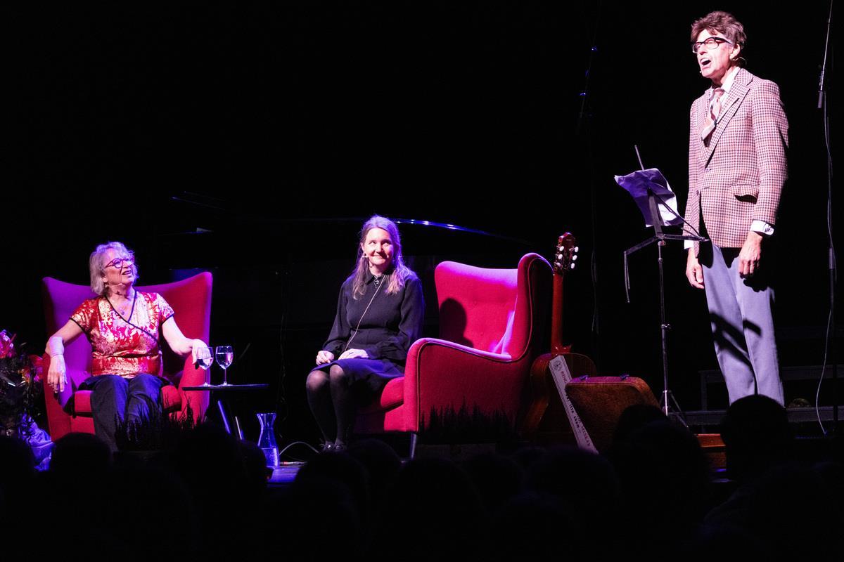 En mann som står på scena mens to damer sitter i hver sin stol. - Klikk for stort bilde