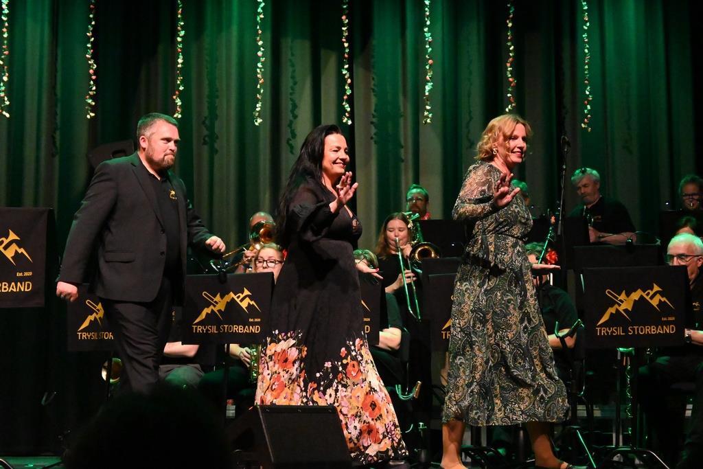 En mann og to kvinner går etter hverandre foran et storband som sitter på en scene - Klikk for stort bilde