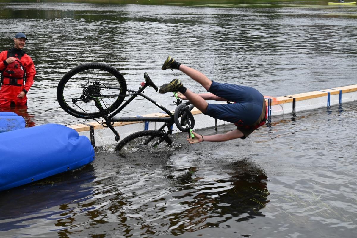 En syklist stuper av sykkelen og uti vannet fra smal planke. En arrangør ser på. - Klikk for stort bilde