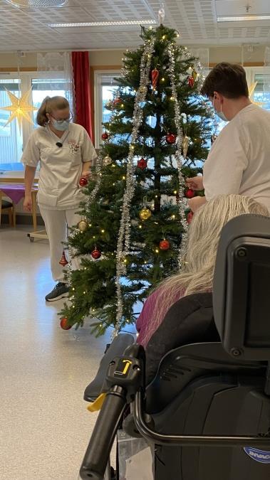 To pleiere pynter juletre mens beboer i rullestol ser på - Klikk for stort bilde